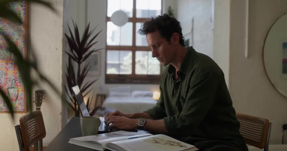 A man reads from a book then types on his laptop
