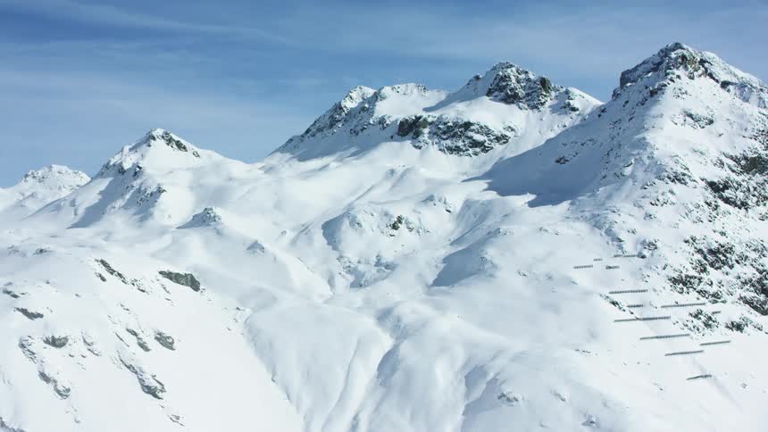 A serene aerial view shows majestic snow covered mountains under a partially cloudy light blue sky