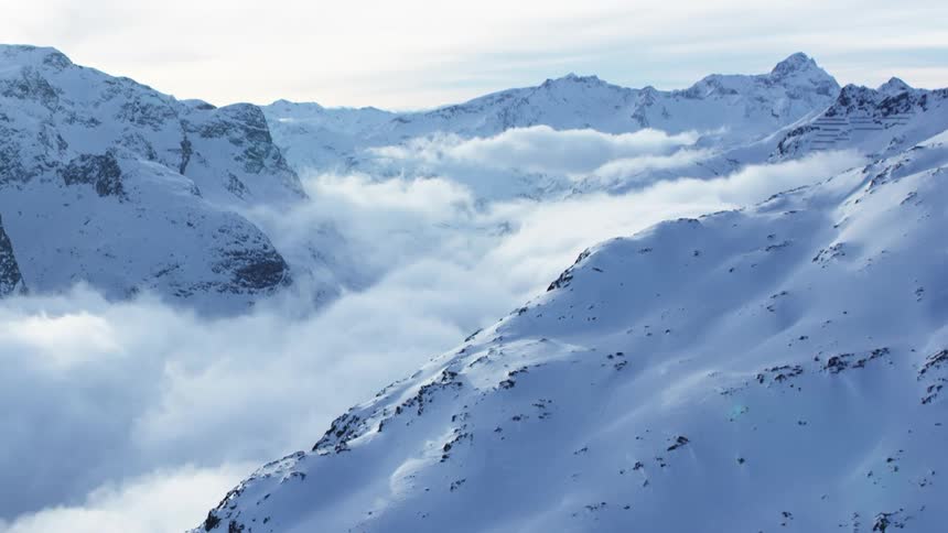 An aerial view reveals a foggy mountain opening with mist drifting between the snowy peaks