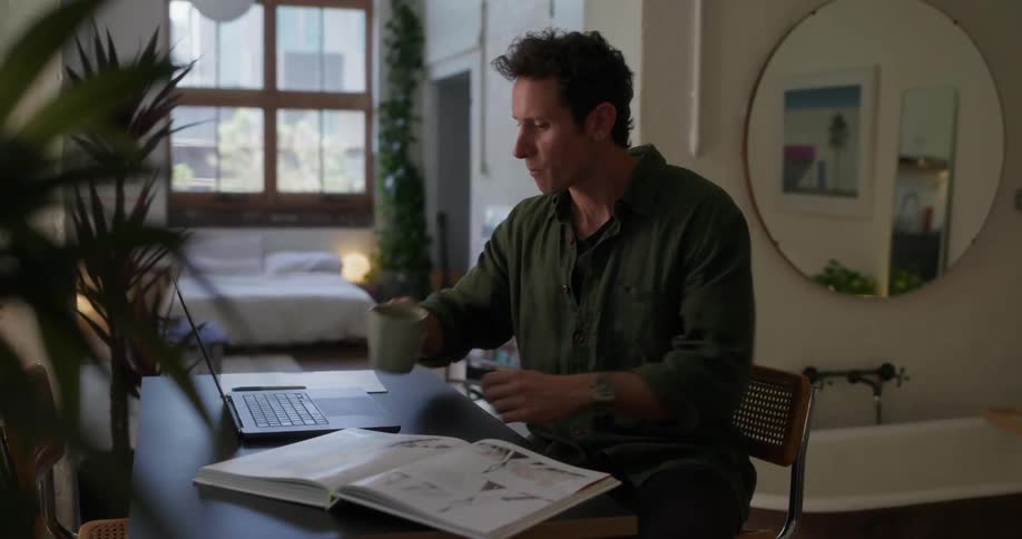 Man sitting at table in modern environment with open book and laptop picking up and drinking from a cup of coffee