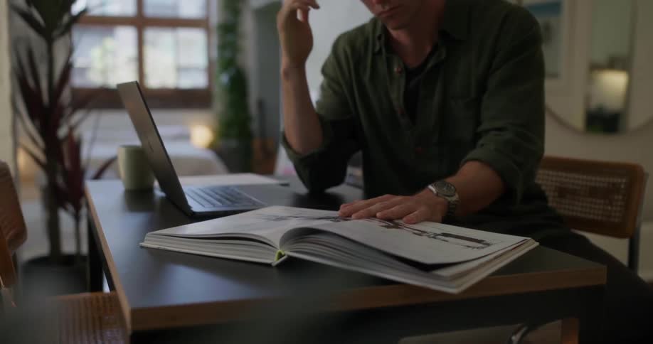 A man in an olive green shirt sits at a desk in a bright modern apartment studying a book with illustrations while holding a pen thoughtfully