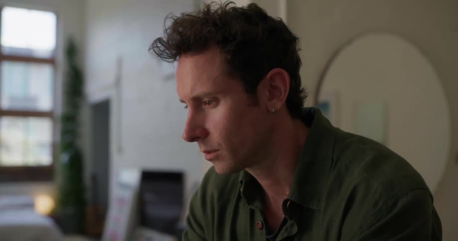 A man in a green shirt sits at a desk types on a laptop and then pauses to think while touching his face