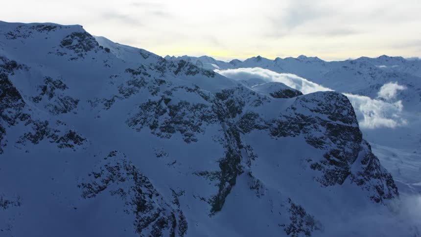 An aerial view of a snowy mountain range reveals rugged ice covered peaks amidst wisps of low hanging clouds