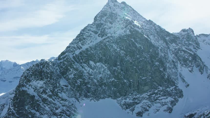 An aerial view captures a towering snowcovered peak with the sun peeking over its summit and a cloudfilled valley below