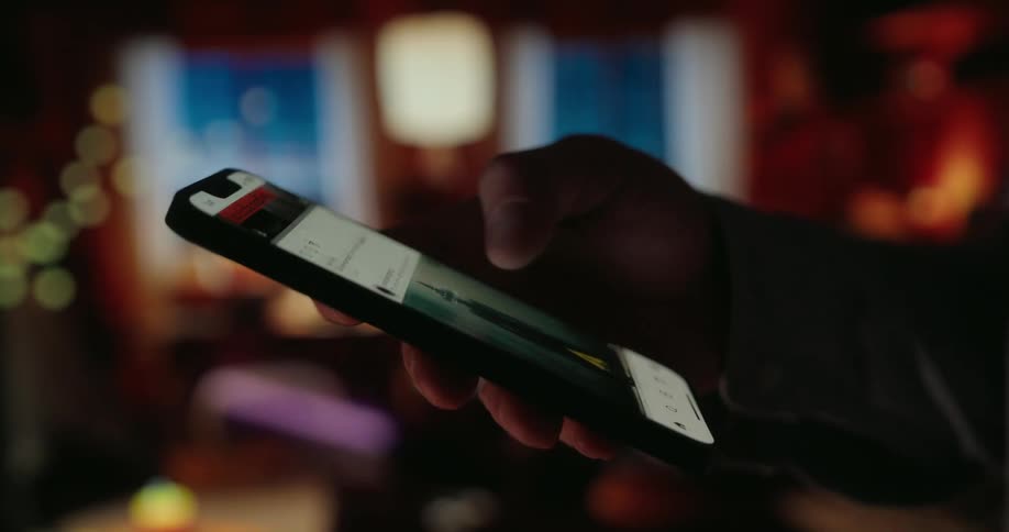 A hand scrolls through content on a smartphone in a dimly lit room with blurred decorations in the background