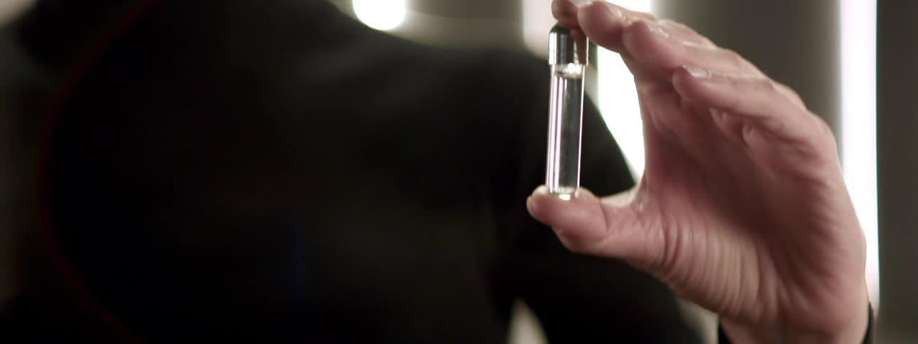 Person wearing black clothing holds two small glass bottles with metal lids one containing a blue liquid and the other a colorless liquid
