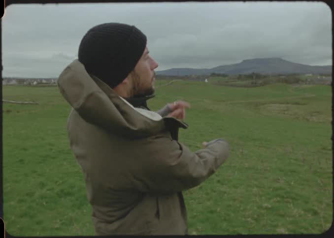 A man in a thick brown jacket stands on a field picks up a red frisbee from the ground and throws it