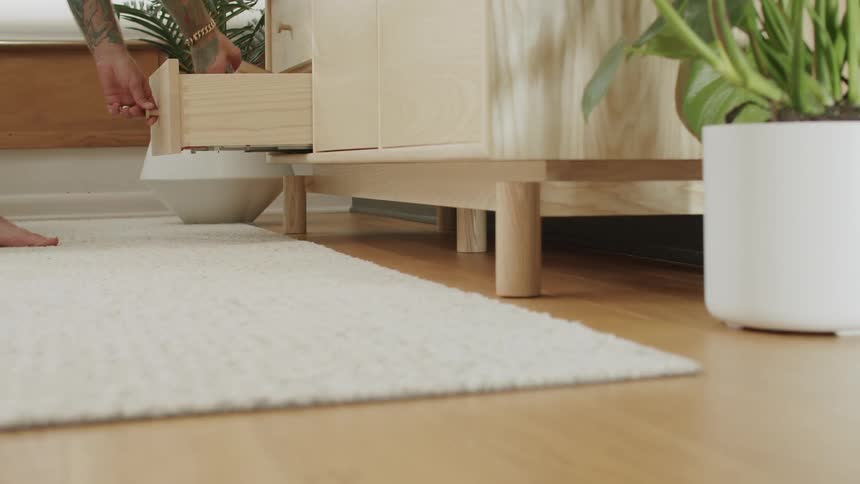 A person is standing barefoot in a living room closing a drawer in a light wooden sideboard