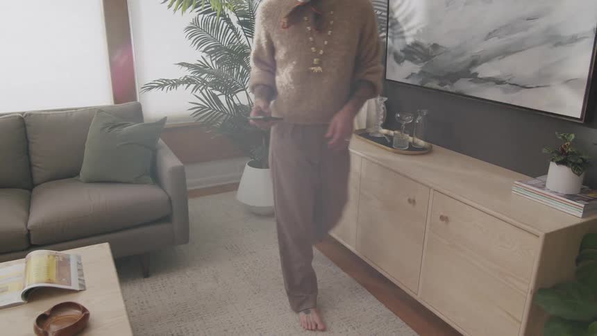 A person is standing barefoot in a living room closing a drawer in a light wooden sideboard next to a grey couch