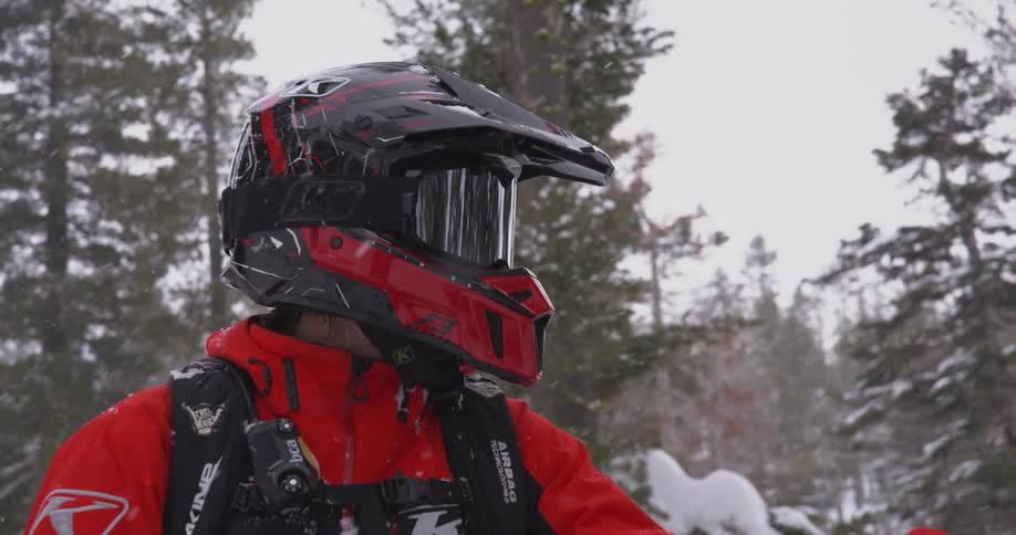 A person wearing a red jacket and a black and red helmet is standing in a snowy forest