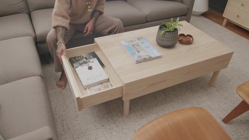 Person putting a book in a drawer attached to a table in the center of a living room