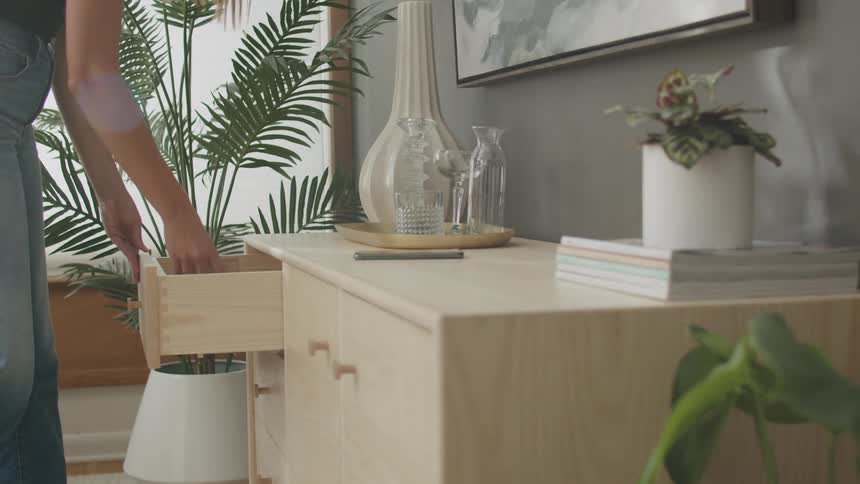 A person in jeans opens a drawer of a light wooden dresser in a room decorated with plants vases and a painting