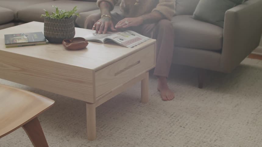 A person sits on a brown sofa barefoot reading a magazine over a wooden coffee table