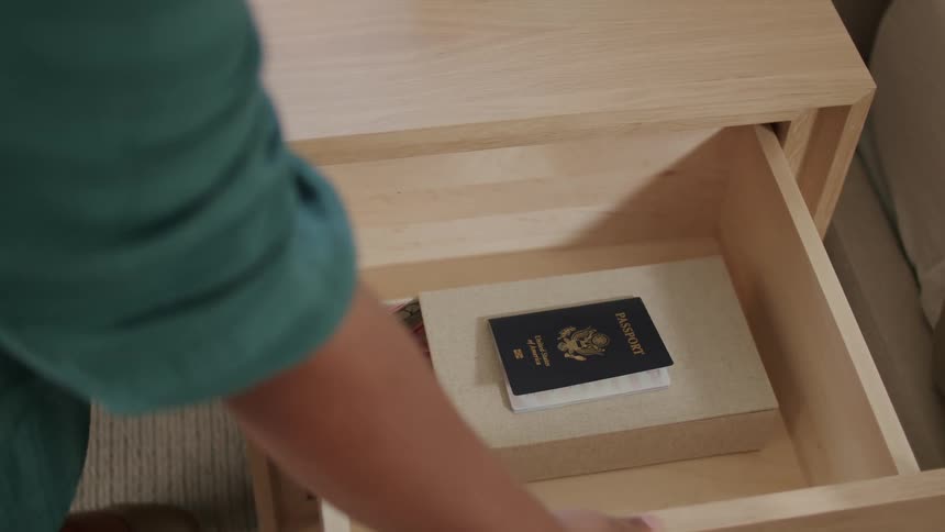 A person opening the drawer of a wooden rack that is next to a bed and taking a passport that is inside