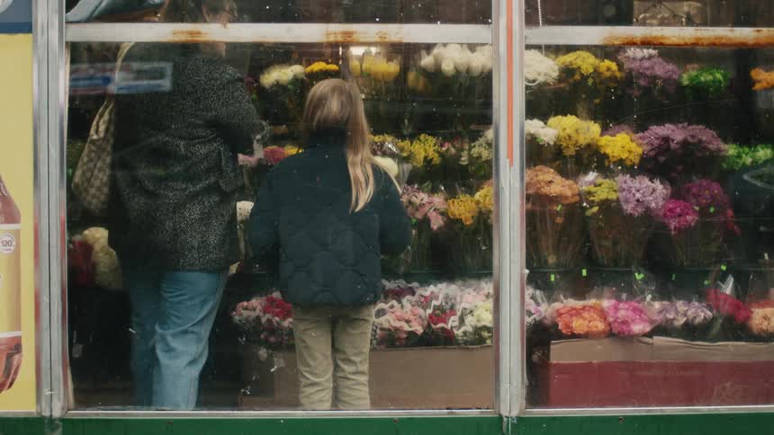 An adult person and a little girl are in an indoor location seen through a window where there are several colors on a shelf and of several different colors