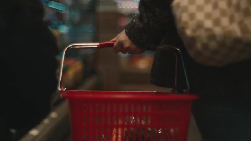 A woman with a checkered purse and gray jacket carries a red shopping basket placing groceries into it