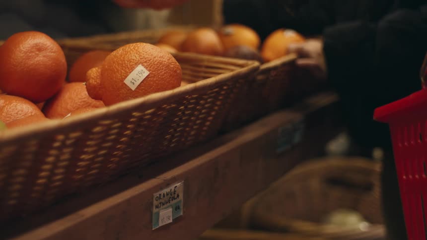 A hand is picking up an orange from a wicker basket in a grocery store