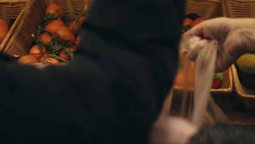 A person taking some oranges from a basket at a market and putting them in a plastic bag and tying it
