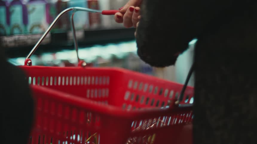 A person carrying a red shopping basket and putting some products inside