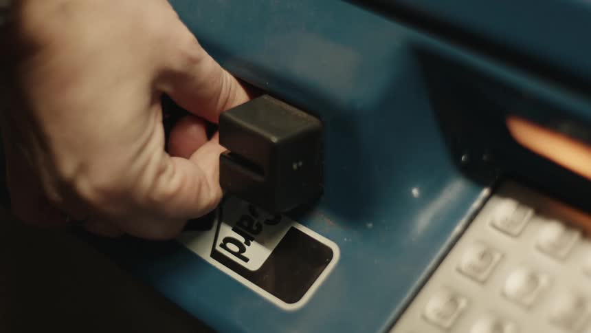 A hand is inserting a gold colored credit or debit card into a blue card reader machine which has a keypad visible to the right