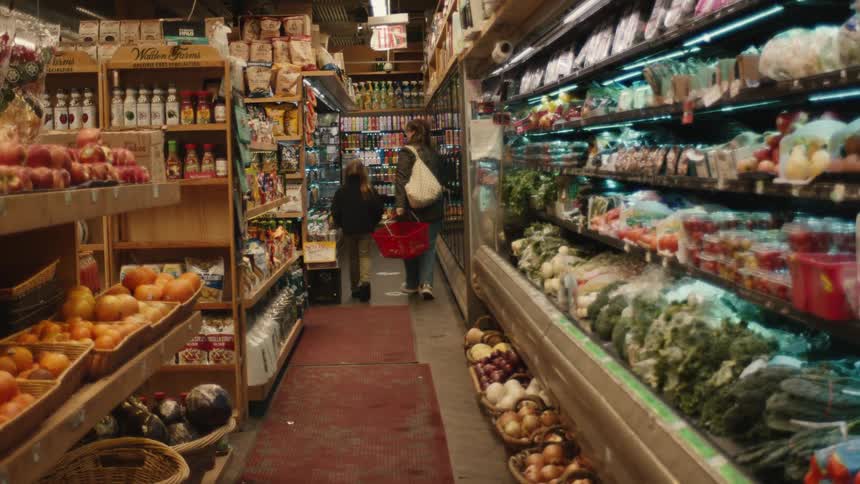 A woman and a little girl are walking down an aisle in a small supermarket and the woman puts some groceries in her basket
