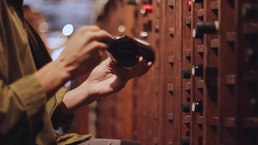 A person in a brown jacket places wine bottles into shelves in a cellar with out of focus people behind them
