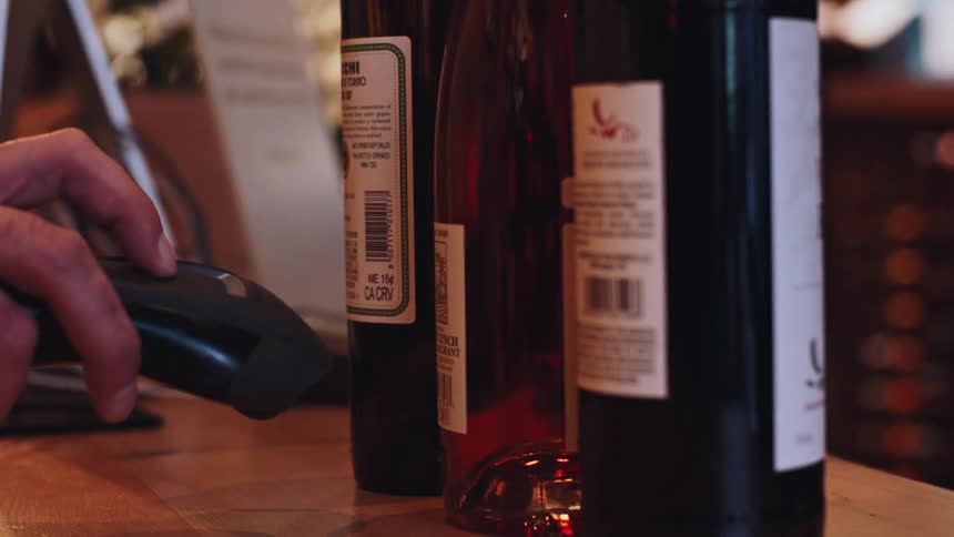 A person is using a barcode scanner to scan the barcode on a wine bottle label with other wine bottles visible in the background