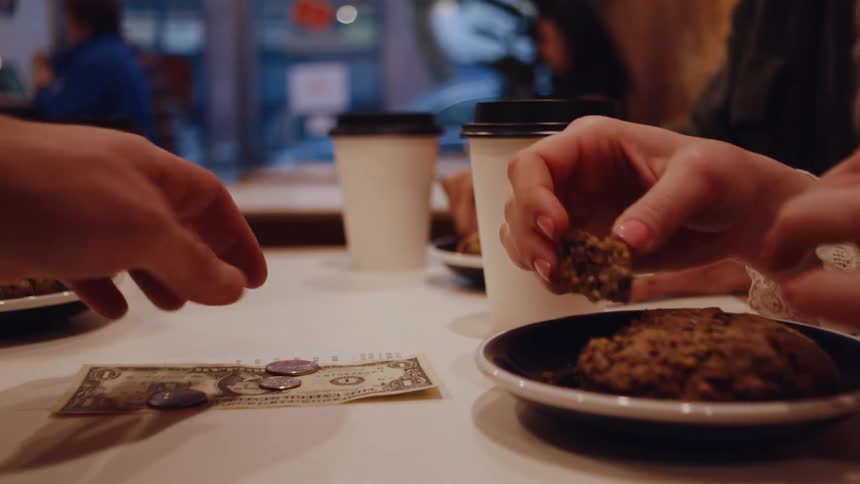Two hands at a caf table one holding a halfeaten cookie and the other reaching for money with coffee cups in the background