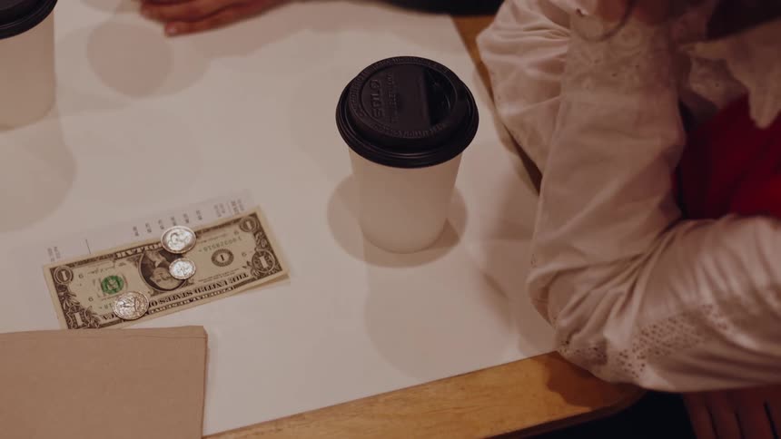 Coffee table with a disposable cup a napkin a tip consisting of a dollar bill and some coins and a person in a lacesleeved blouse sitting next to it