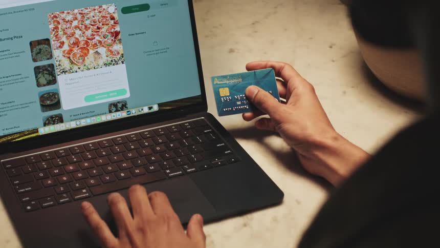 A person types on a macbook air to order pizza online holding a credit card in one hand