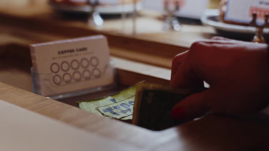 A hand places a bill into a wooden tip drawer next to a stack of coffee cards
