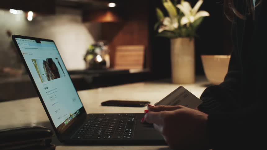 Person holds a credit card while making an online purchase on a laptop on the kitchen table