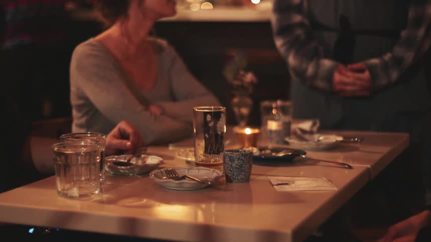 Three people are seated at a table in a dimly lit restaurant with drinks and dishes in front of them while a server holds a bill being paid by one of the customers