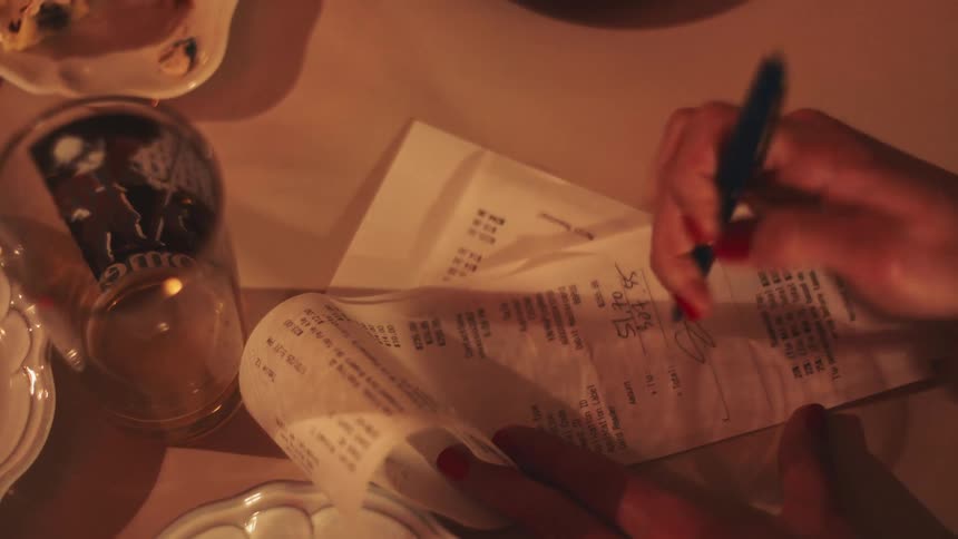 A hand with redpainted nails holds a long receipt and writes on it with a blue pen at a restaurant table