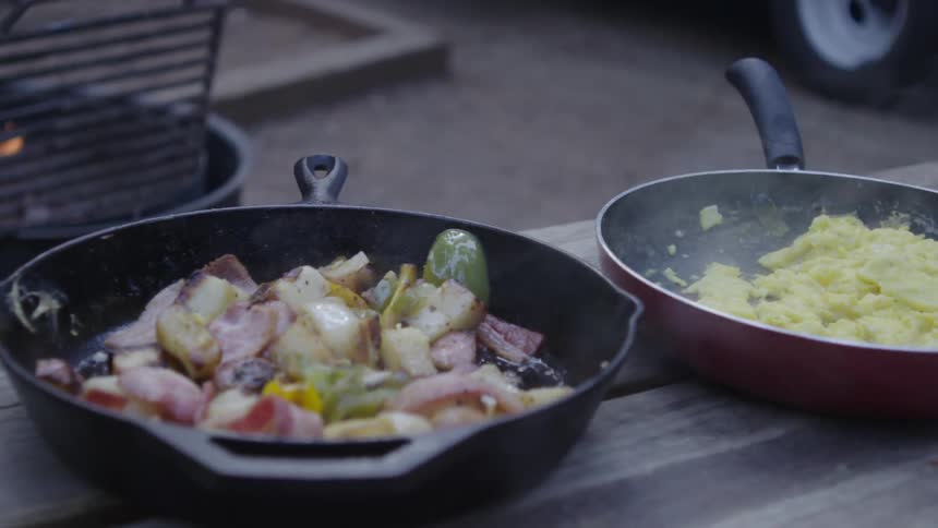 Two skillets of food on a picnic table with a fire pit in the background