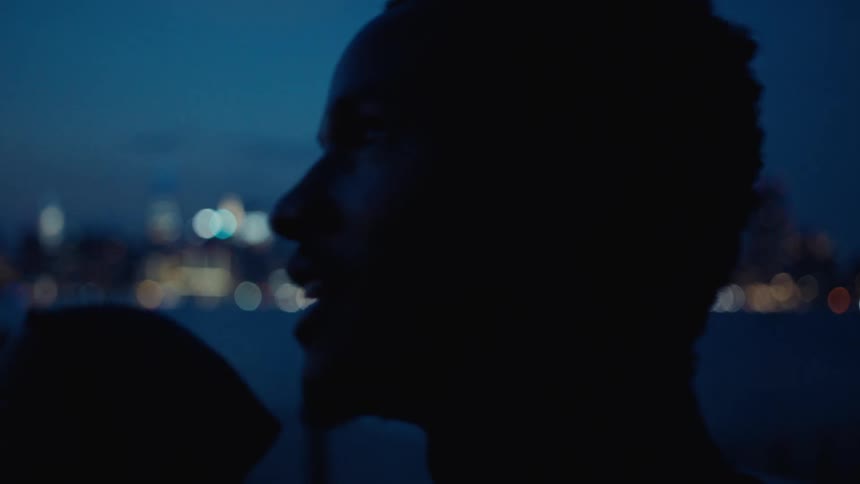 Man talking to his friend looks up at the sky while standing on the waterfront across from the city skyline at night