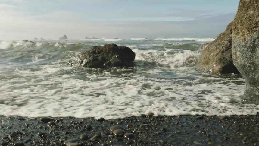 Ocean waves crash against large rocks near the shore under a partly cloudy sky