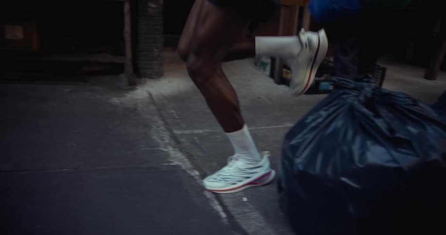 A person wearing white socks and red sneakers jumps over large black garbage bags on a sidewalk while jogging