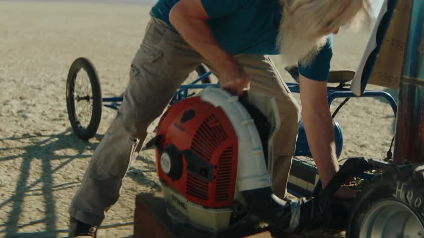 An elderly bearded man is working on his vehicle while in the desert on a sunny day