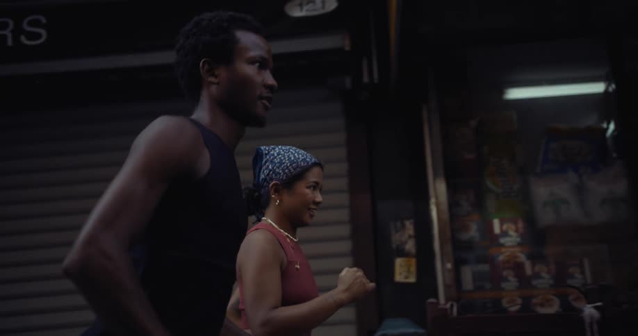 A man and woman are running in slow motion past a shop in a neighborhood