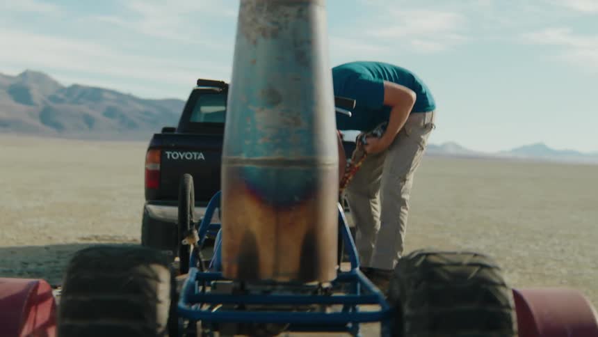 Man unchains a cart loaded on a trailer towed by a pickup truck that is parked in the desert