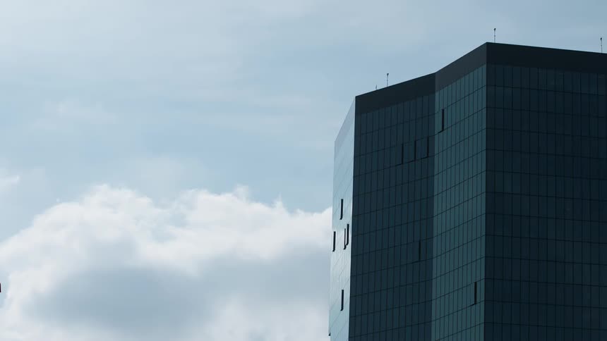 A modern dark colored skyscraper with a geometric facade stands against a light blue sky with fluffy white clouds