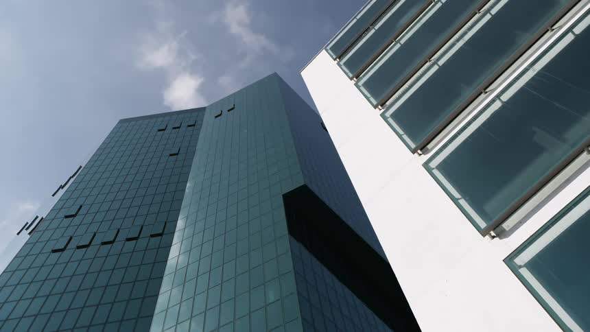 A modern building with a dark facade and small rectangular windows juts into a partly cloudy sky contrasted by a white building with large rectangular windows on the right