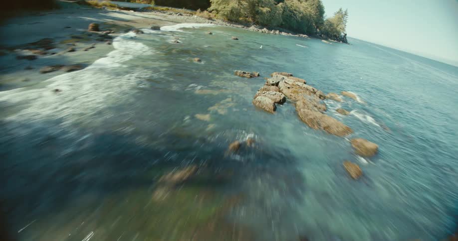 Sea waves crash against submerged rocks near the coast with a forest in the background
