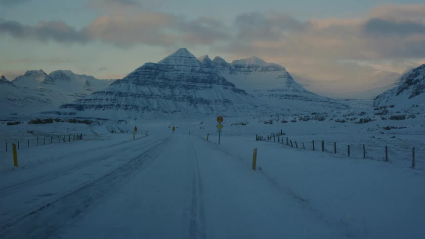 A snow covered road stretches towards majestic layered mountains under a cloudy sky