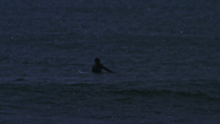 A person is floating on a surfboard in the ocean at dusk with only the silhouette visible against the dark water