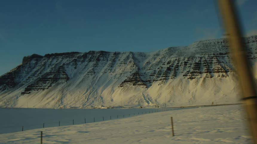 Snowcovered mountains with layered textures rise above a flat icy foreground under a clear sky