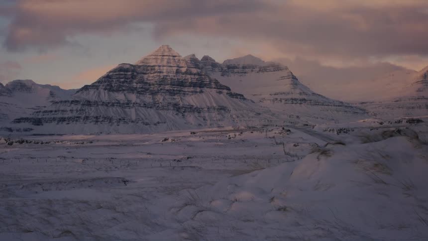 A snowcovered landscape stretches out under a cloudy sky with a distinctive pyramidal mountain in the distance