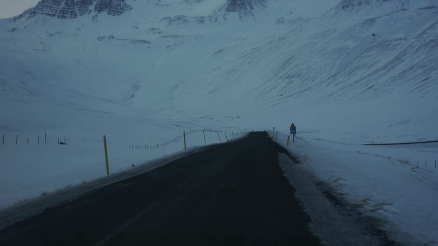 A dark asphalt road cuts through a snowy mountainous landscape under a dim overcast sky