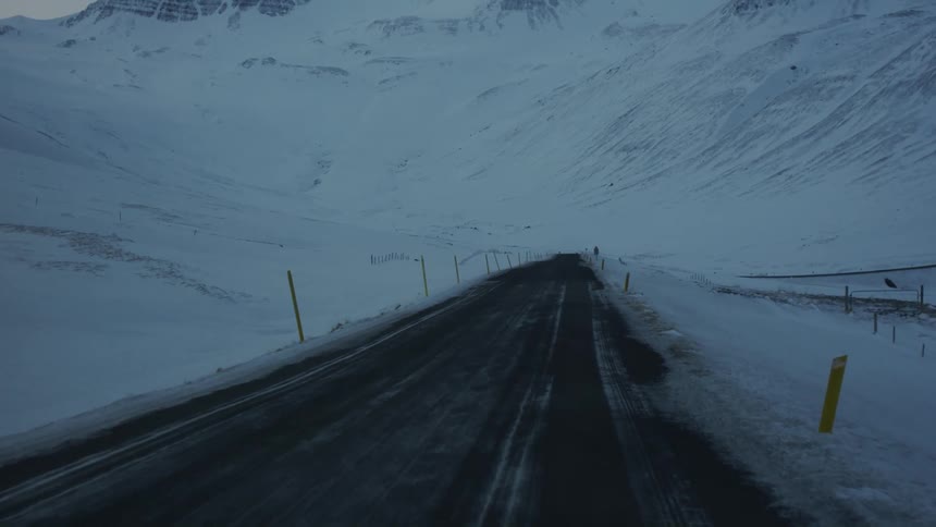 A car travels on a snowy road through a mountain range on a cloudy day